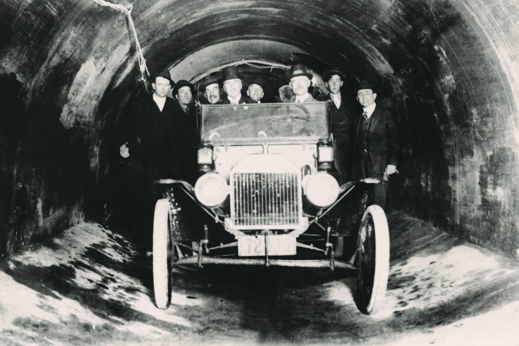 Photograph shows nine men in a tunnel who are standing in and behind a 1915 convertible car. 