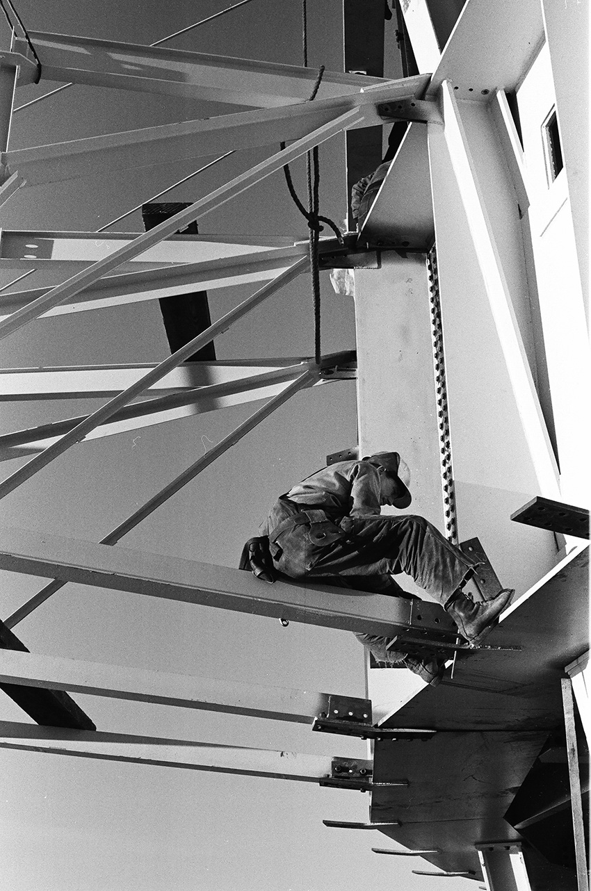The photograph shows a man sitting on a long, thin piece of steel that is perpendicular to a tall steel structure. 