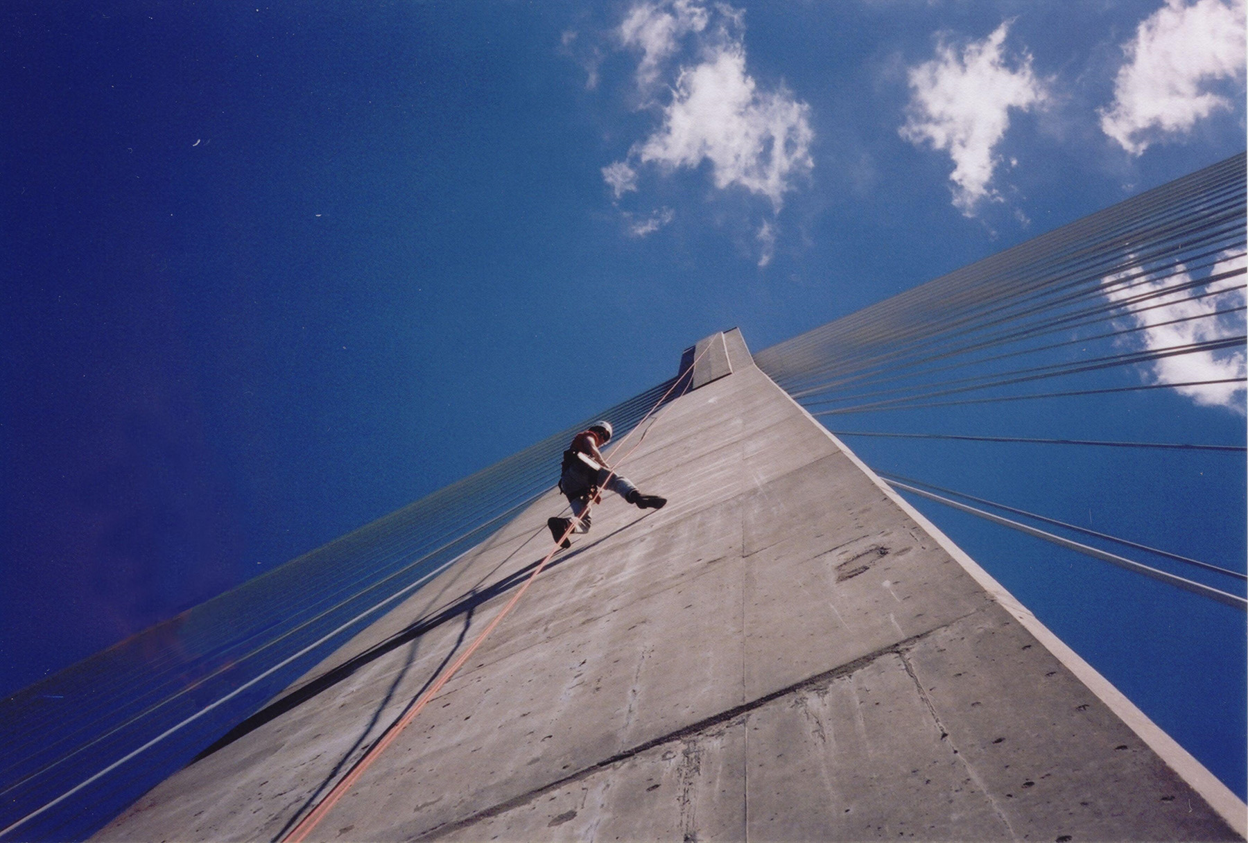 A ground-level picture shows a person rope climbing down the side of a cable bridge. 
