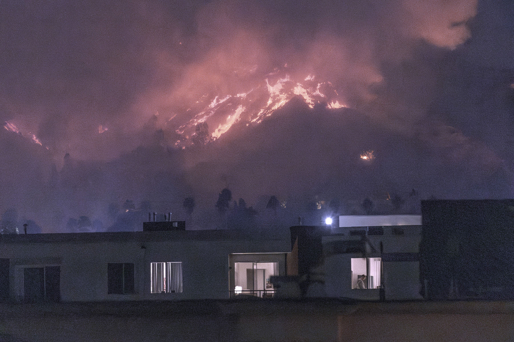 One-level buildings are shown against the backdrop of a burning hillside. 