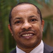 Man with brown skin in jacket and tie smiles at the camera. He has a mustache.